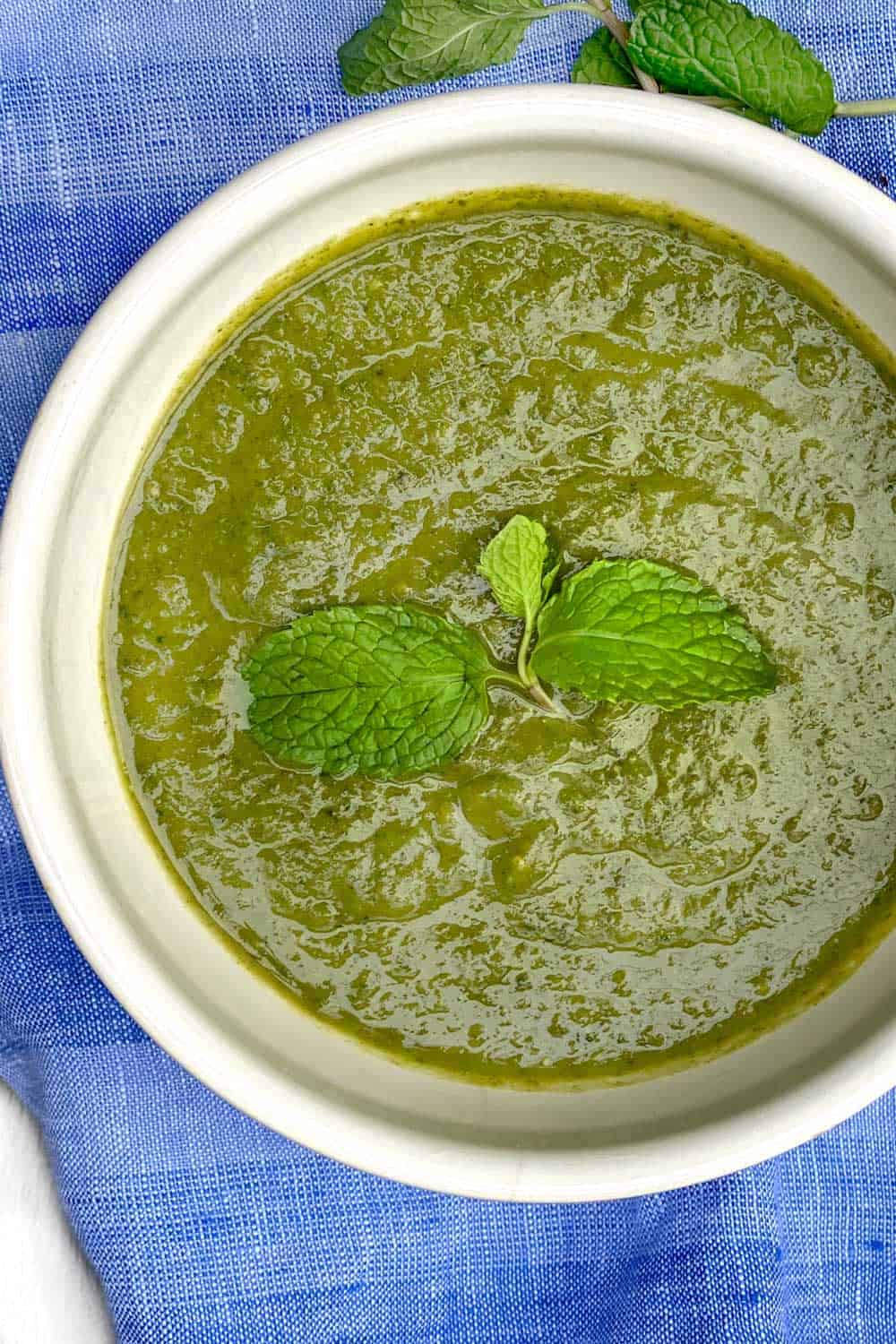 close up overhead looking into a white bowl on a vivid blue dish towel, bowl is filled with bright green sweet pea and spinach soup with a sprig of fresh mint in the middle.