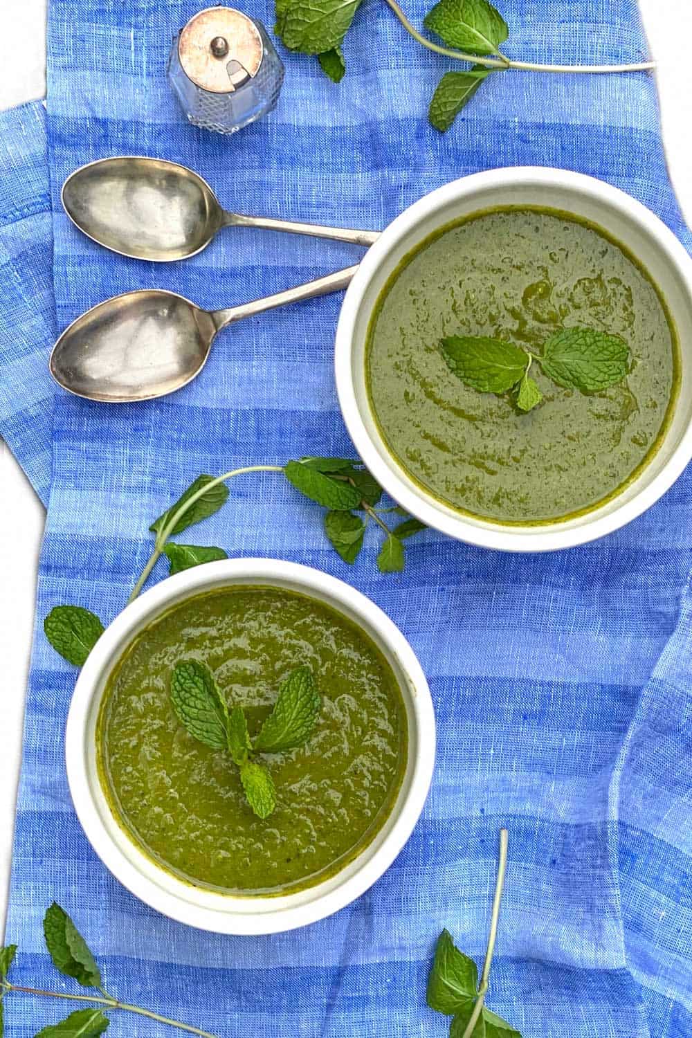 overhead shot of two white bowls, on a vivid blue dish towel, filled with vivid green pea and spinach soup. Fresh mint sprigs garnish the soup and are scattered around the dish towel, and there are two soup spoons