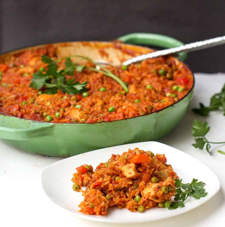 jollof rice in a light green round Dutch oven with a sprig of parsley on top, plus a small white plate with a portion of jollof rice on it