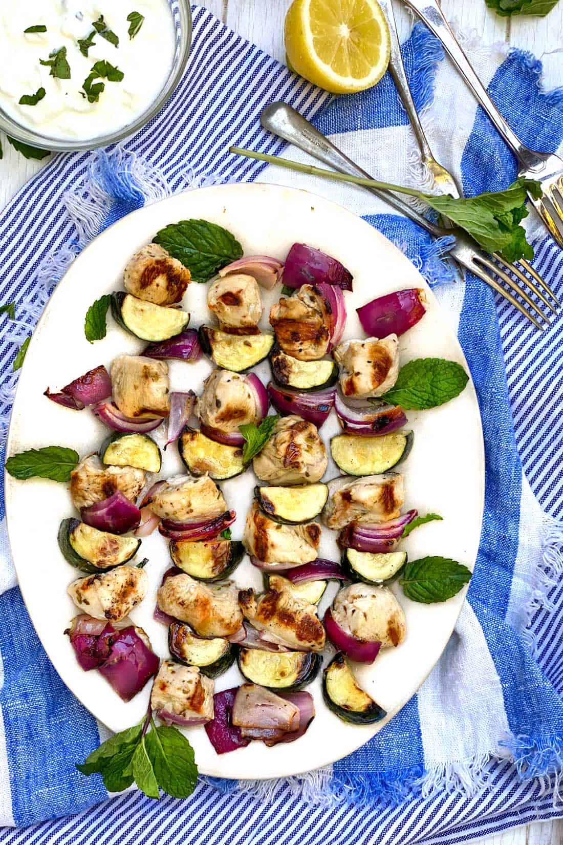 4 chicken souvlaki kebabs with red onions and zucchini, removed from the kebab, on a plate, on a vivid blue and white striped dish towel with a small bowl of tzatziki and a half lemon