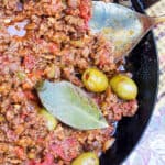 cuban picadillo: ground beef stew with olives, in a black skillet with green olives and a bay leaf