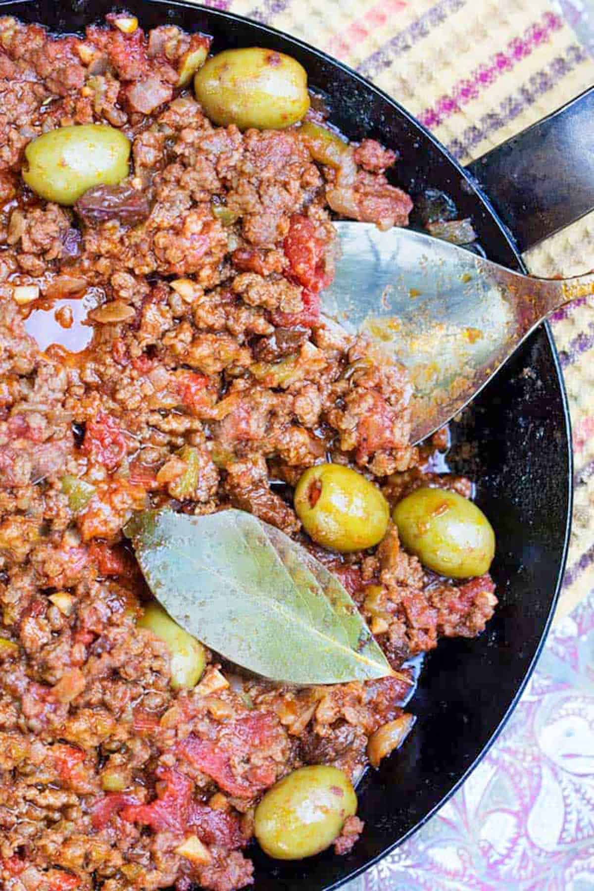 cuban picadillo: ground beef stew with olives, in a black skillet with green olives and a bay leaf