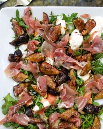 grilled figs, prosciutto and burrata arranged on a bed of arugula on a porcelain platter.