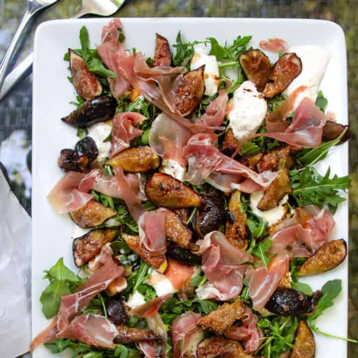 grilled figs, prosciutto and burrata arranged on a bed of arugula on a porcelain platter.