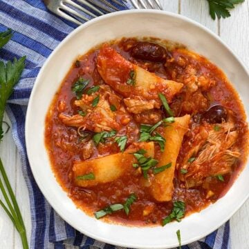 white bowl on a blue striped dish towel, filled with Greek chicken stew with potatoes and olives in tomato sauce