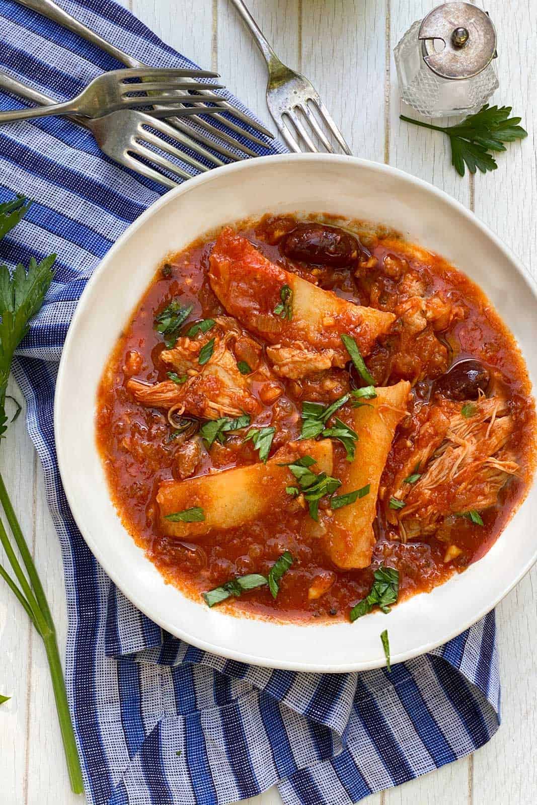 white bowl on a blue striped dish towel, filled with Greek chicken stew with potatoes and olives in tomato sauce