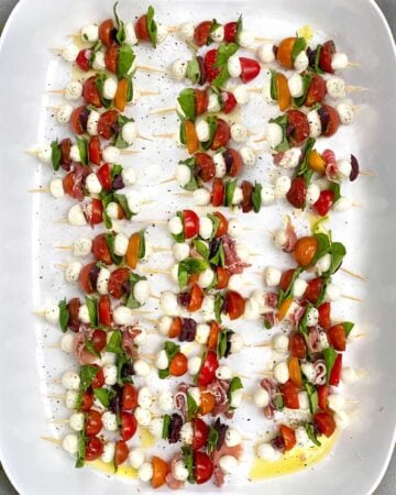 3 dozen mini caprese skewers on a white tray, with tomatoes, basil leaves and mini mozzarella balls