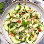 Thai cucumber salad with sliced red chili peppers, chopped mint leaves and chopped peanuts, in a white bowl on a pin-striped black and white dish towel with 3 forks