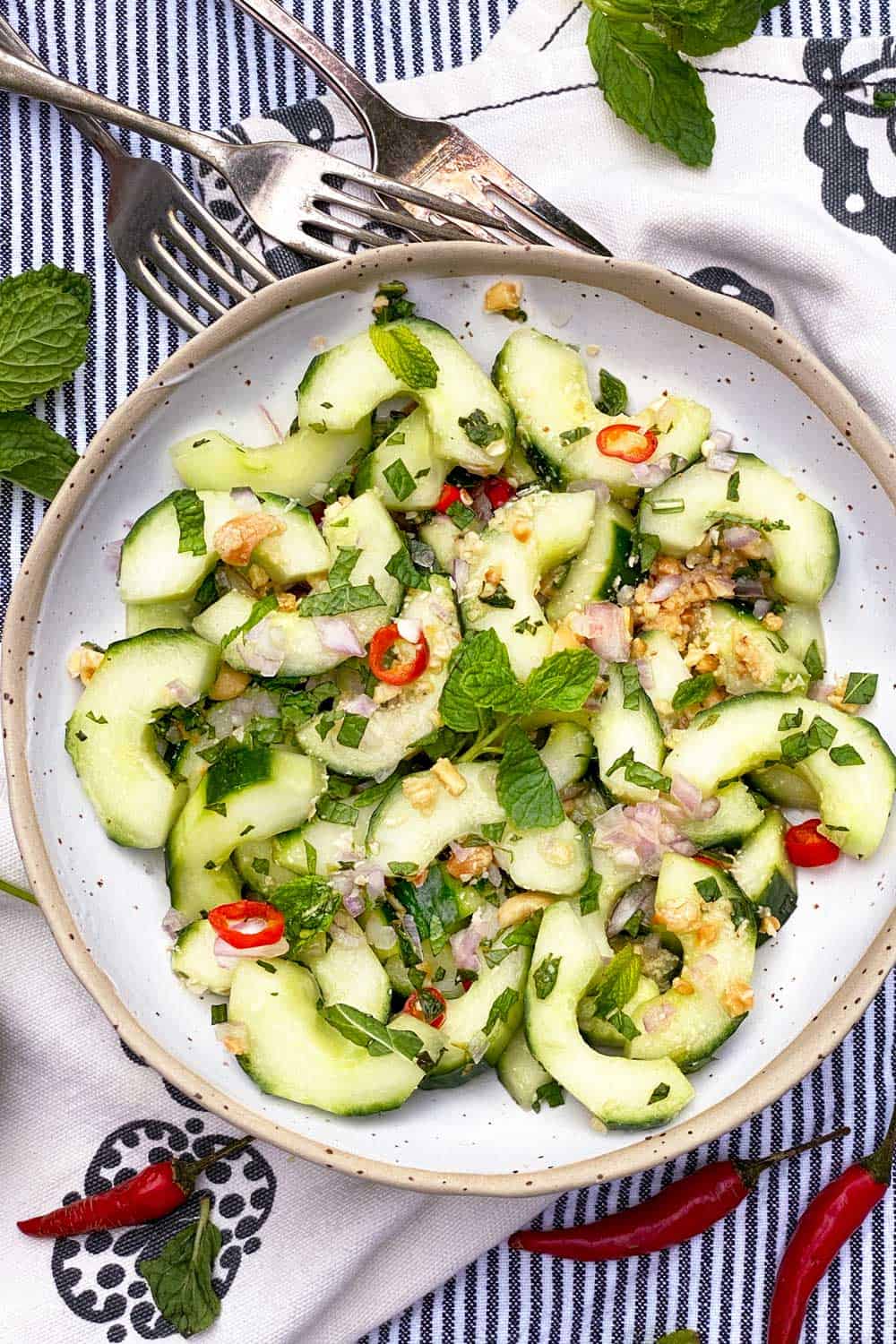 Thai cucumber salad with sliced red chili peppers, chopped mint leaves and chopped peanuts, in a white bowl on a pin-striped black and white dish towel with 3 forks