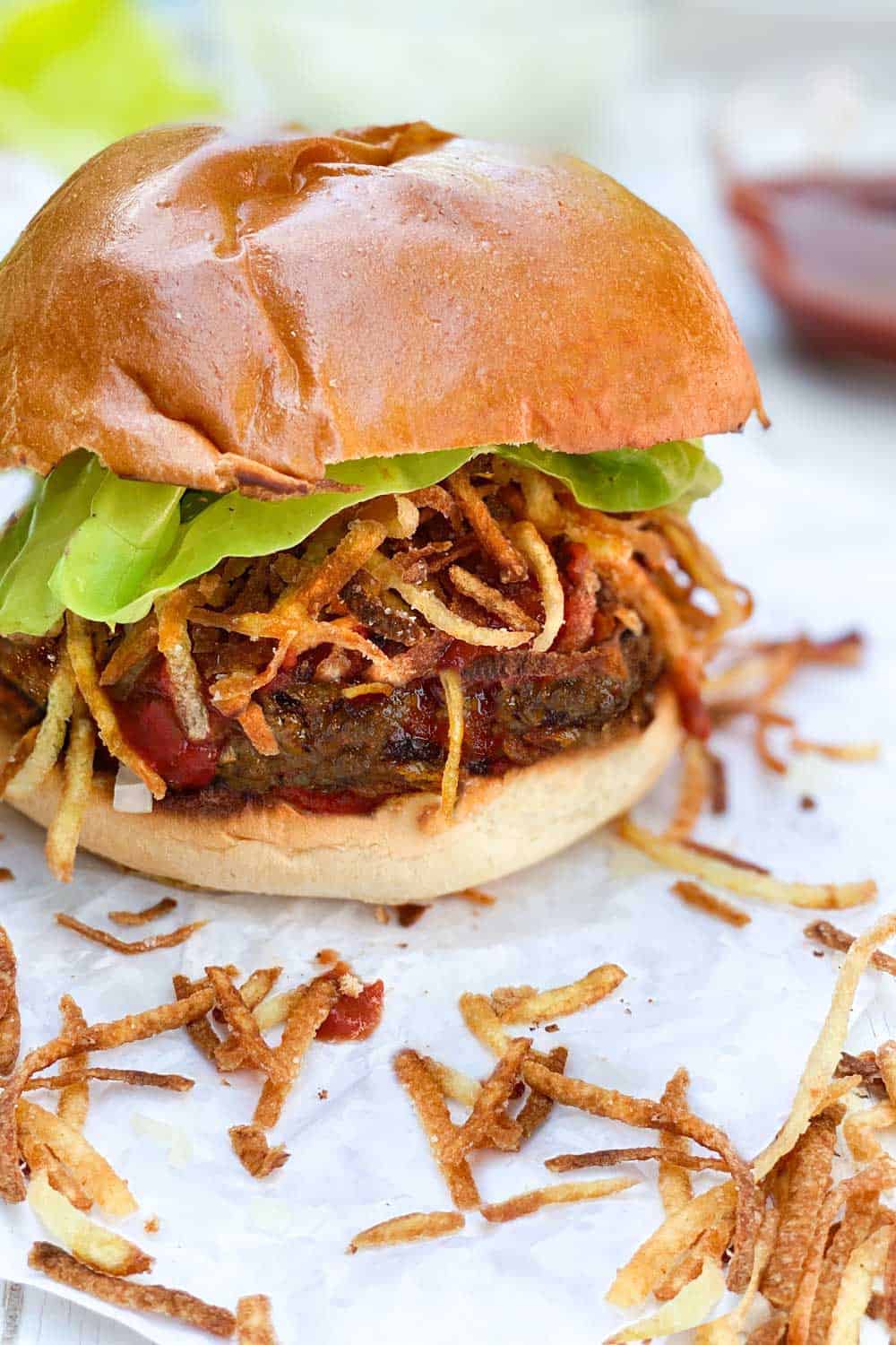 A Cuban frita burger topped with shoestring fries and a lettuce leaf, in a bun, on a piece of white parchment