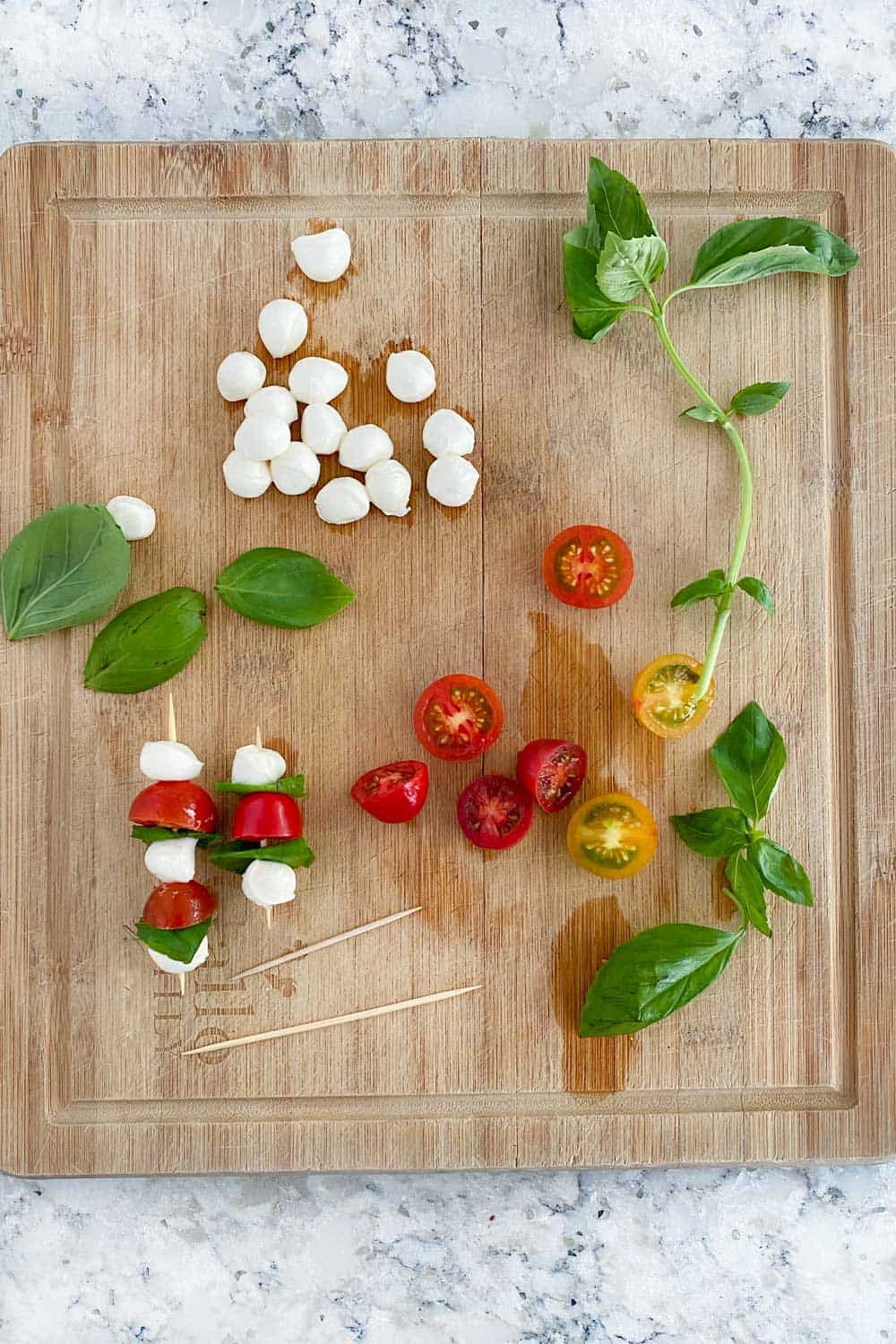 wooden cutting board with ingredients for making mini caprese skewers: a dozen mini mozzarella balls, a handful of basil leaves, a few halved cherry tomatoes and a few toothpick skewers