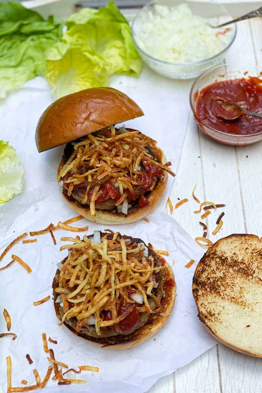 how to make the frita cuban, showing two burger patties on two buns with chopped onions and fried potatoes and special sauce on top, lettuce leaves on the side and the bun tops on the side.