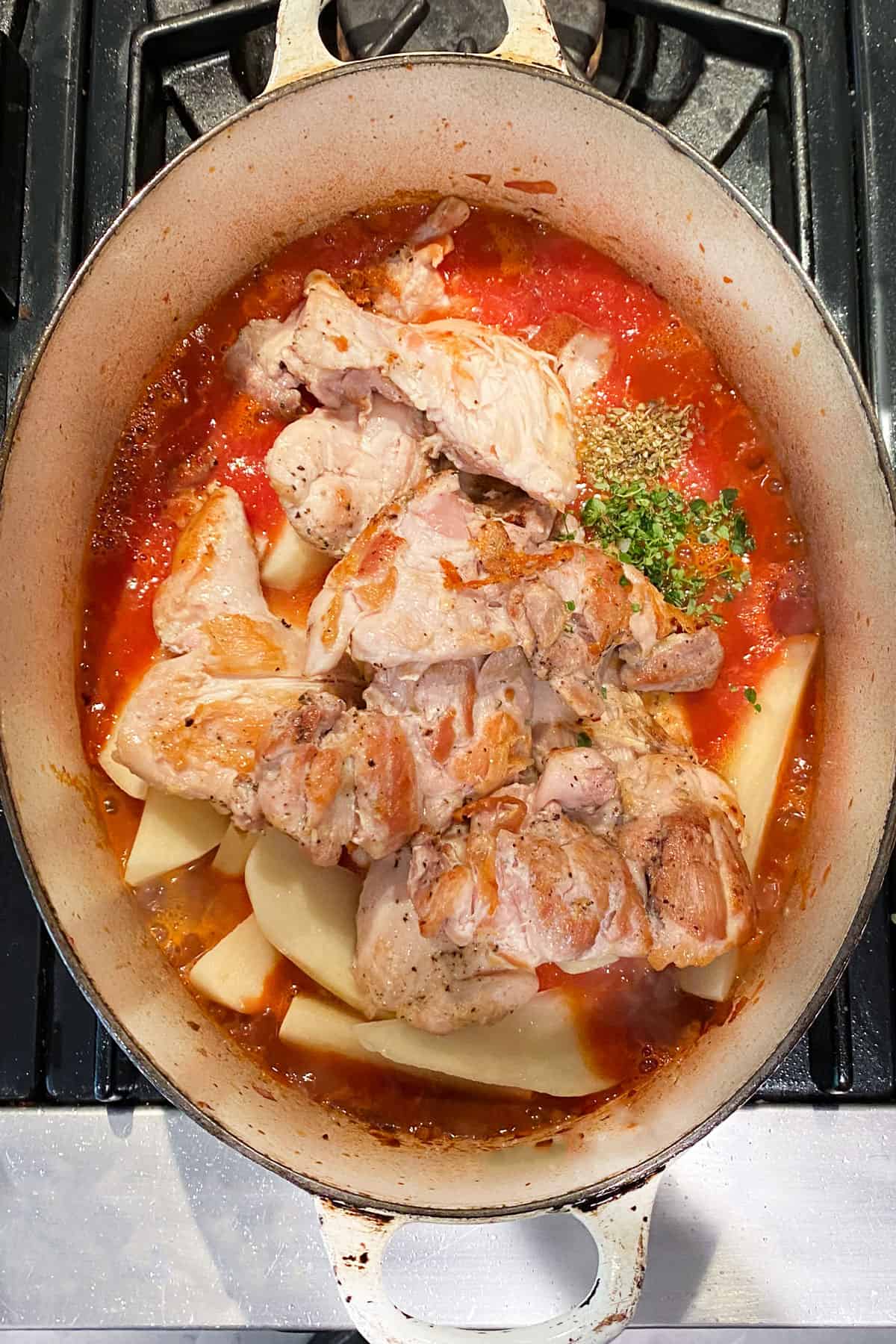 Oval dutch oven filled with browned chicken thighs, raw potato wedges, tomato sauce and spices.