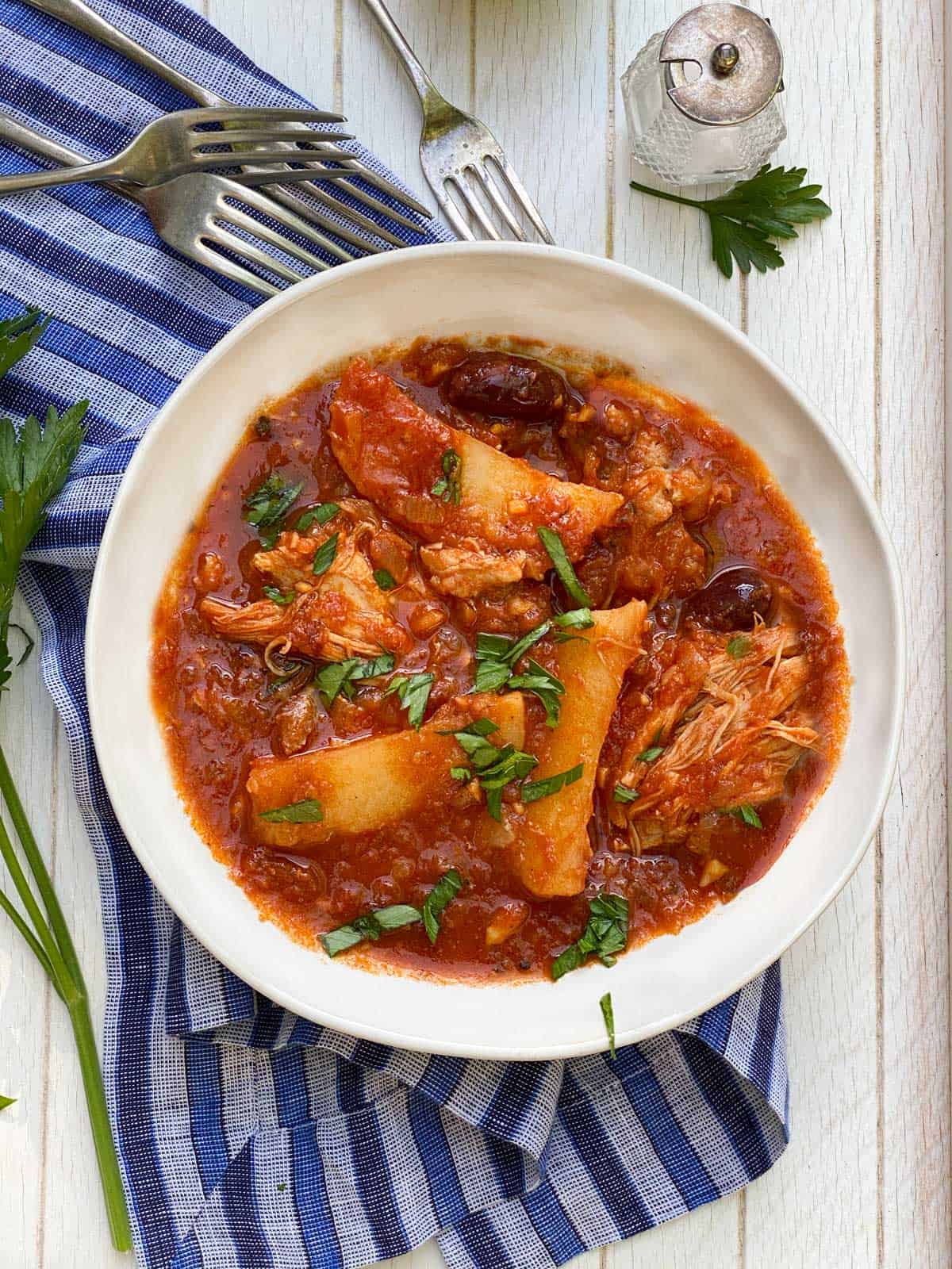 white bowl on a blue striped dish towel, filled with Greek chicken stew with potatoes and olives in tomato sauce