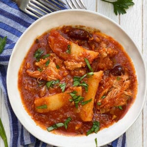 white bowl on a blue striped dish towel, filled with tender chicken stew with potato wedges and olives in tomato sauce