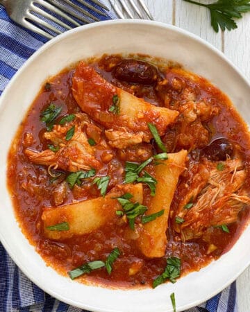 white bowl on a blue striped dish towel, filled with tender chicken stew with potato wedges and olives in tomato sauce