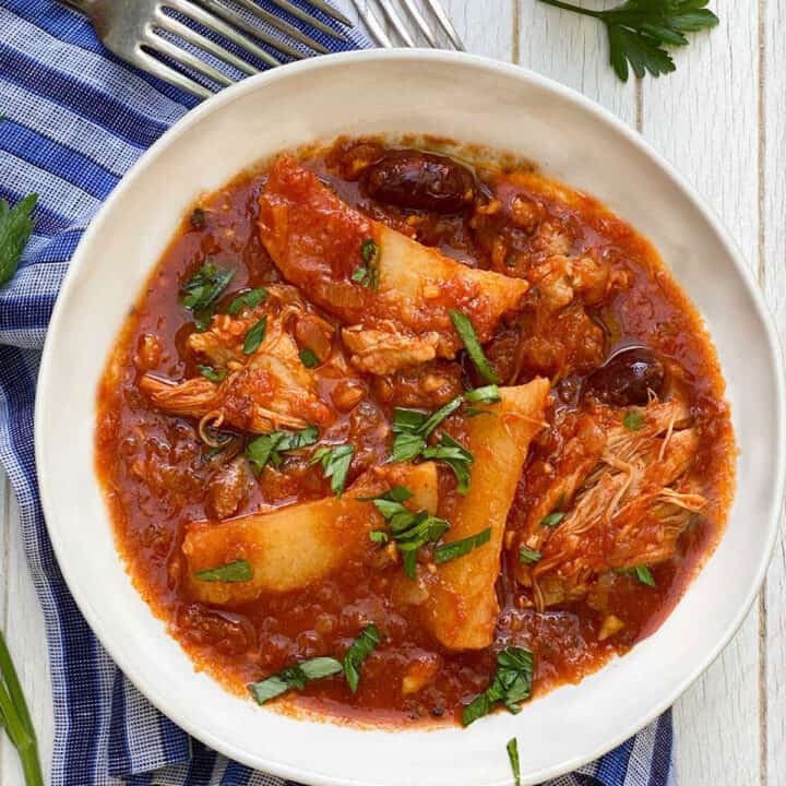 white bowl on a blue striped dish towel, filled with tender chicken stew with potato wedges and olives in tomato sauce