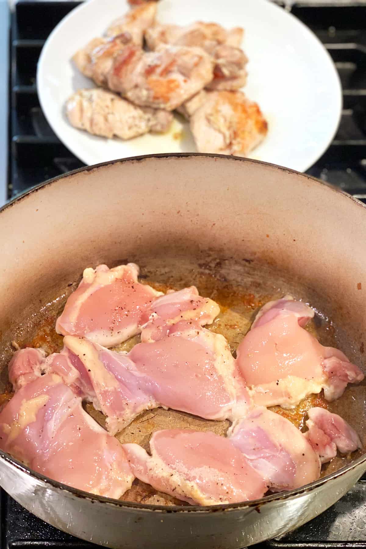 several boneless chicken thighs browning in an oval Dutch oven with a plate in the background holding a few pieces of browned chicken thighs.