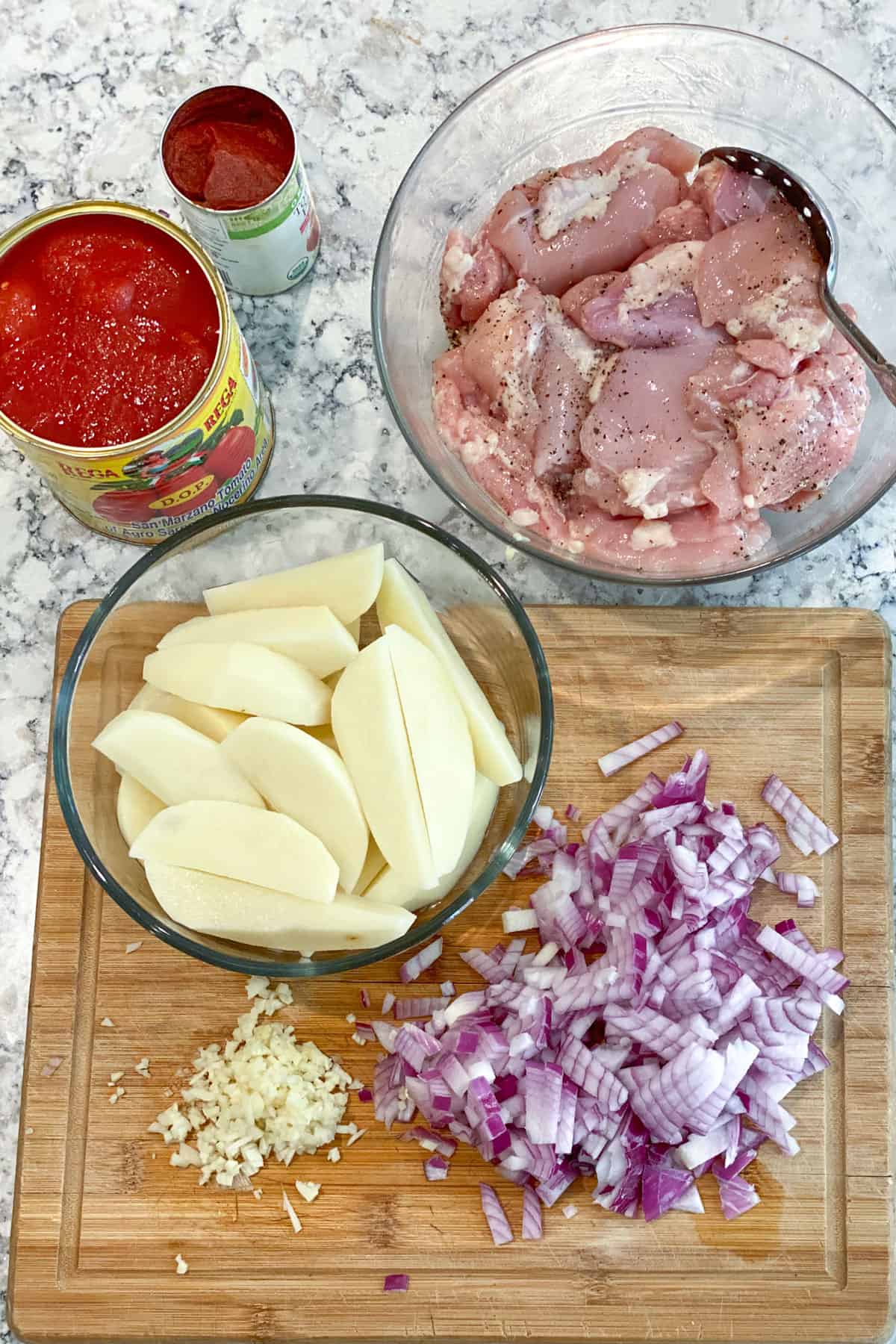 Prepped ingredients on a countertop: Potato wedges in a glass bowl, Chopped red onion in a bowl, chopped garlic on a wooden cutting board, canned tomatoes opened, boneless chicken thighs salted and peppered in a bowl.