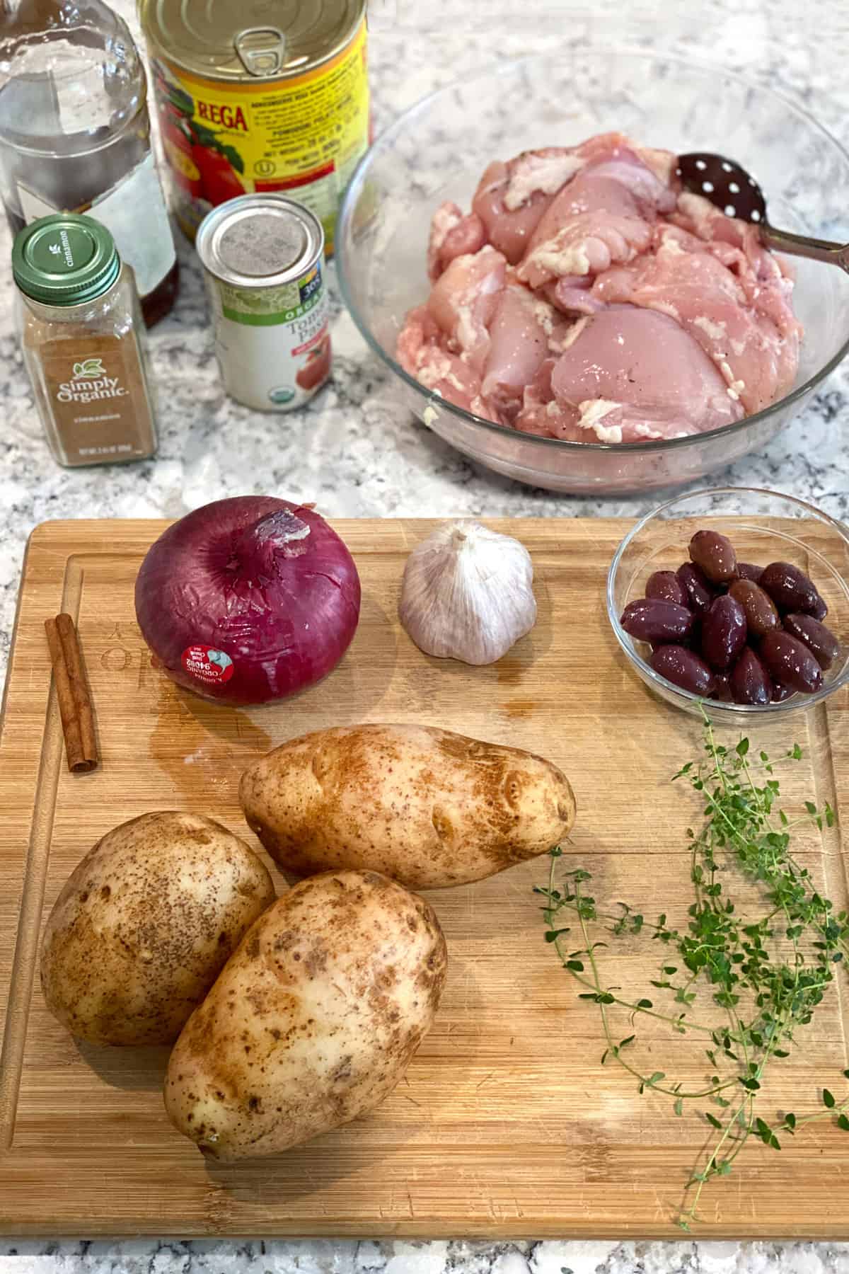 Ingredients on a granite countertop: glass bowl of raw boneless chicken thighs, 3 russet potatoes, 1 red onion, a few sprigs of thyme, a small bowl of kalamata olives, a cinnamon stick, a head of garlic, can of tomato paste, can of plum tomatoes and jar of cinnamon.