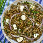 a white bowl filled with lentil salad and cubes of feta, on a blue striped dish towel with some fresh chives on the side and a couple of pink chive flowers