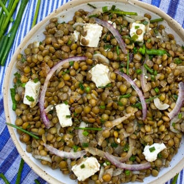 a white bowl filled with lentil salad and cubes of feta, on a blue striped dish towel with some fresh chives on the side and a couple of pink chive flowers