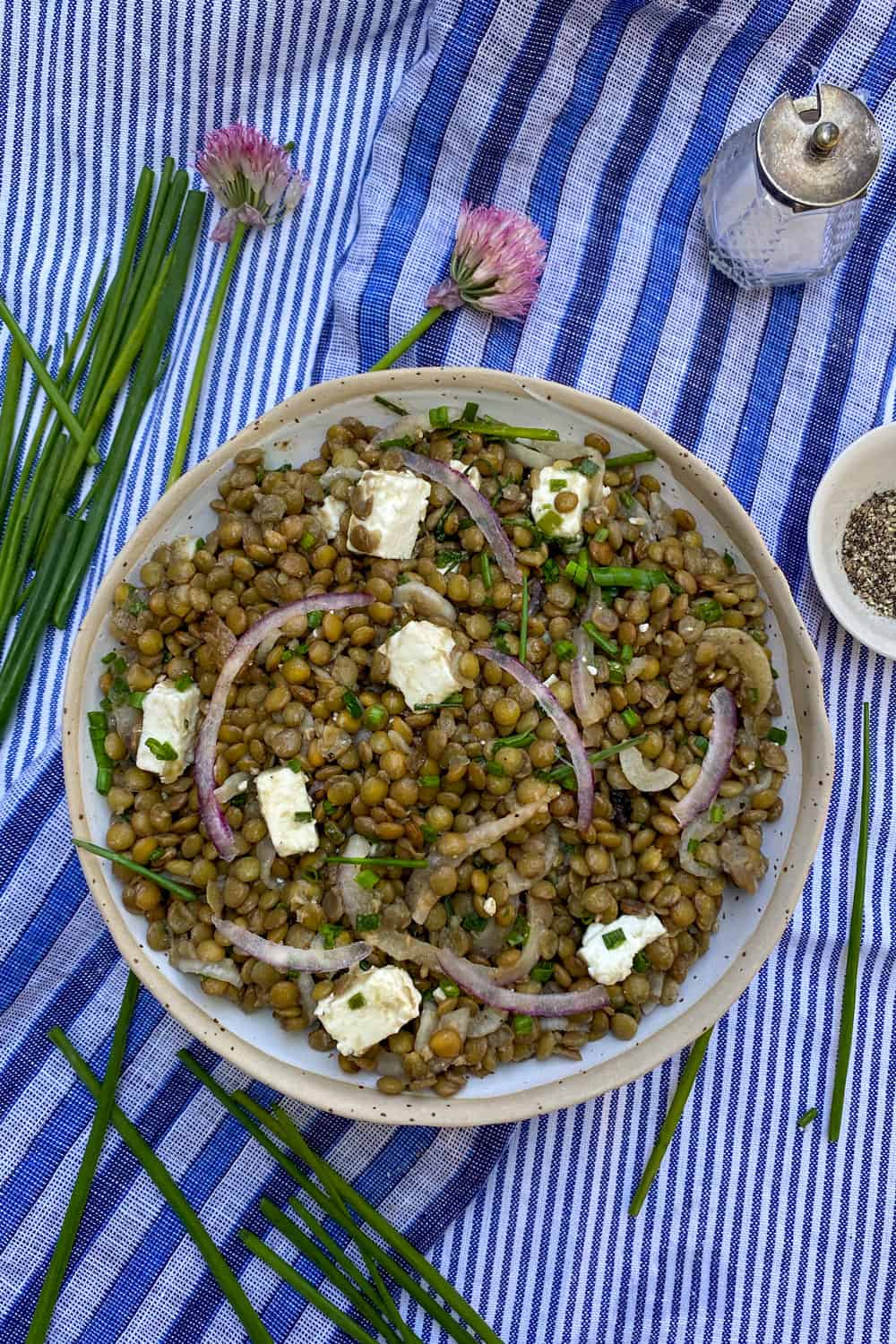 a white bowl filled with lentil salad and cubes of feta, on a blue striped dish towel with some fresh chives on the side and a couple of pink chive flowers