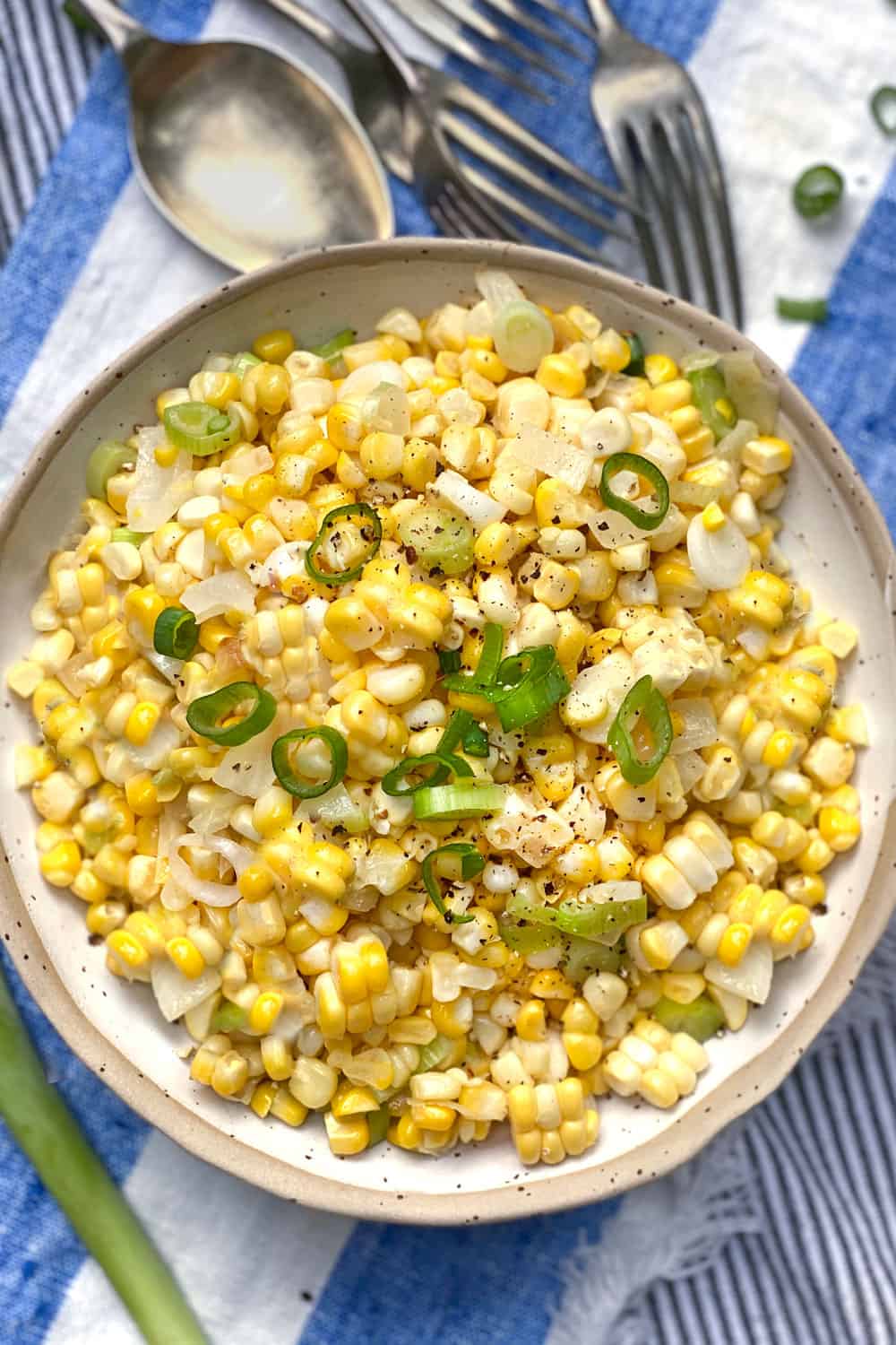 white bowl filled with sauteed corn and scallions, a blue and white striped cloth napkin underneath and several forks and a spoon on the side.