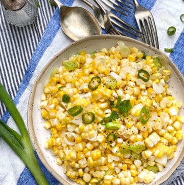 a white bowl filled with sautéed corn kernels and chopped scallions. The bowl sits on a blue and white striped cloth napkin and several pieces of flatware are in the background