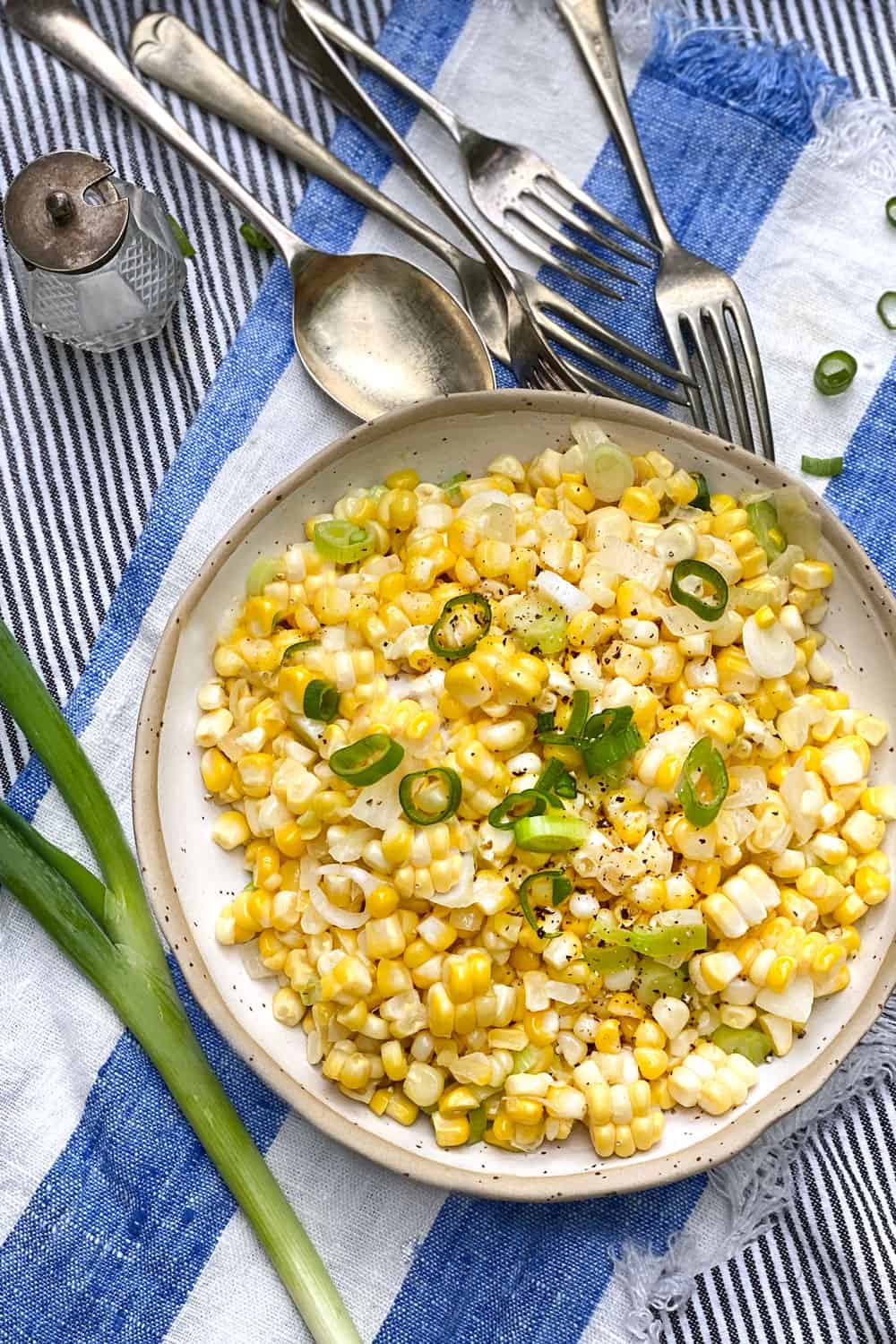 a white bowl filled with sautéed corn kernels and chopped scallions. The bowl sits on a blue and white striped cloth napkin and several pieces of flatware are in the background