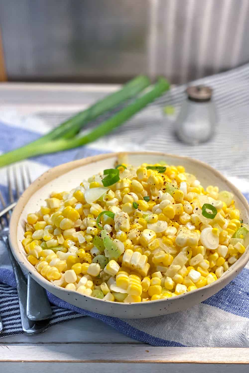 a white bowl filled with sautéed corn kernels and chopped scallions, in the background is a whole scallion and a small glass salt shaker.