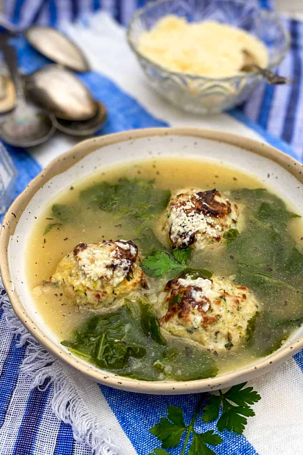 a white bowl on a blue and white striped dish towel, filled with Italian wedding soup, broth, 3 turkey meatballs and wilted kale, with a pile of spoons and a small bowl of parmesan in the background