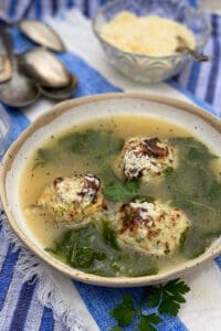 a white bowl filled with Italian wedding soup, broth, 3 turkey meatballs and wilted kale, with a pile of spoons and a small bowl of parmesan in the background