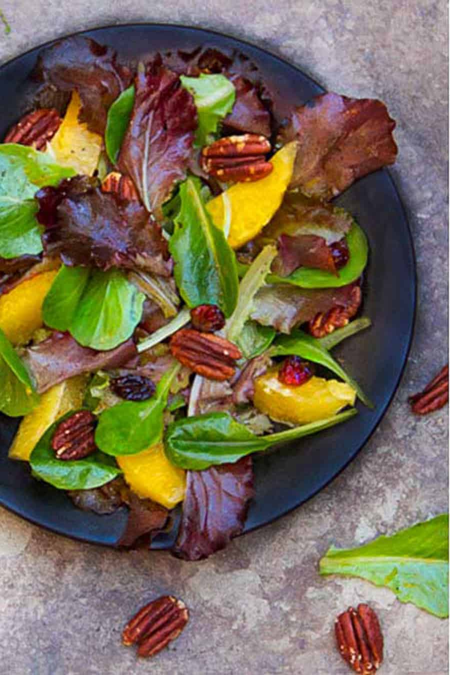 black plate topped with a salad of mixed baby greens, oranges, pecans and cranberries.