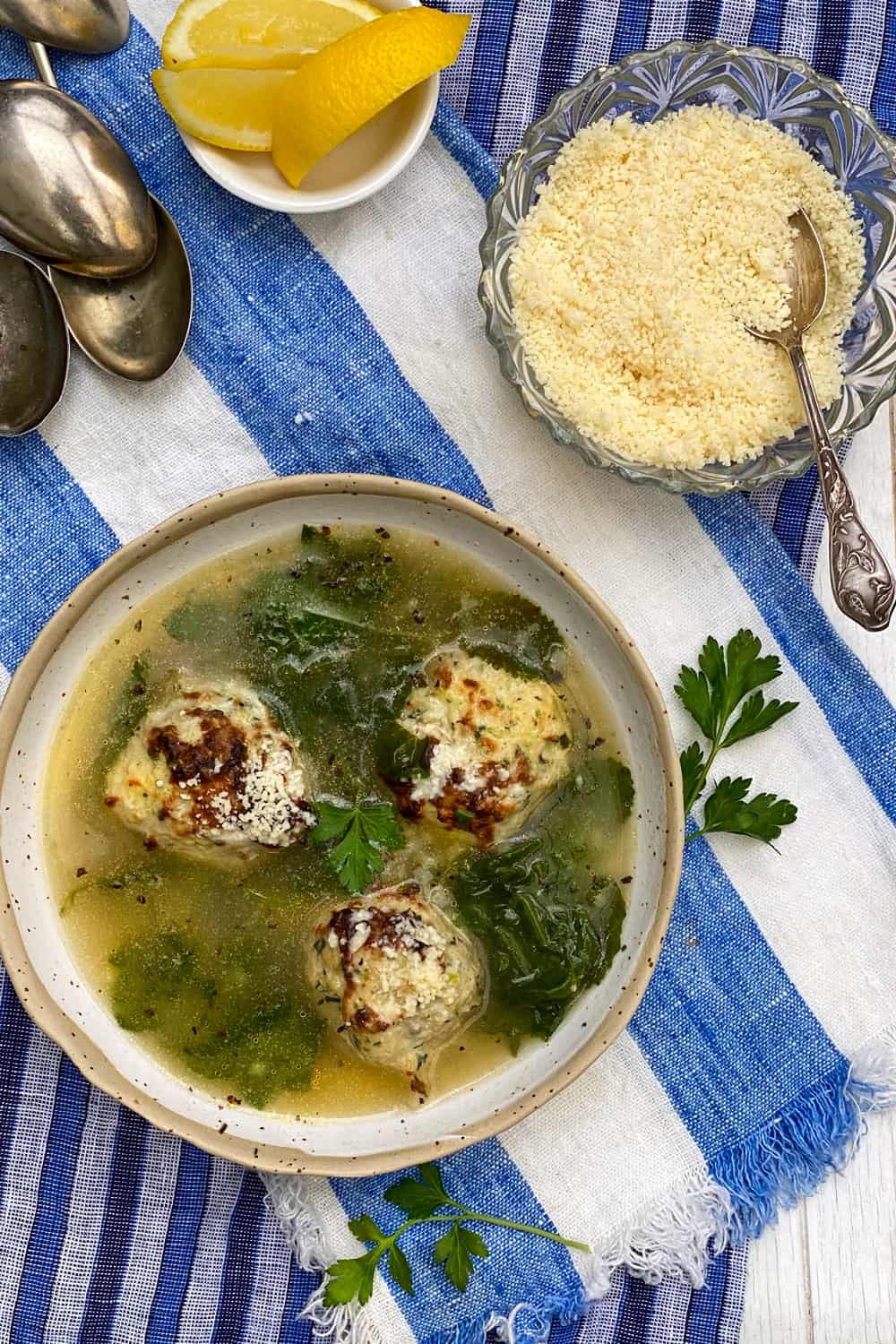a white bowl on a blue and white striped dish towel, filled with Italian wedding soup, broth, 3 turkey meatballs and wilted kale, with a pile of spoons and a small bowl of parmesan in the background