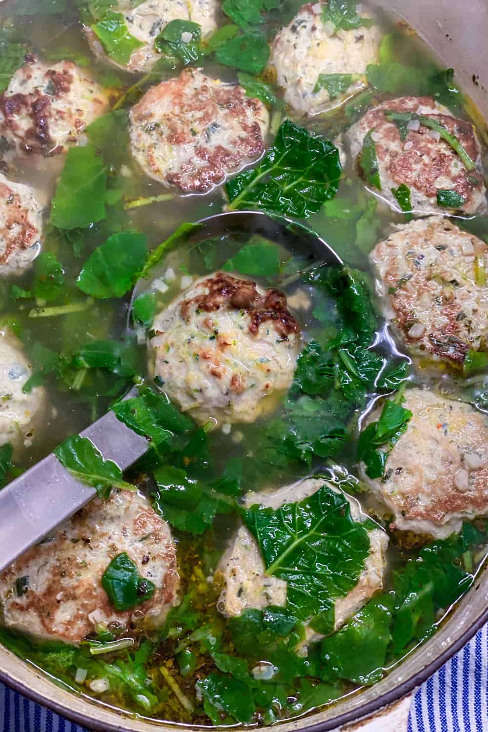 close up looking down into a pot of Italian Wedding soup, showing a ladle with a meatball and greens in it, surrounded by more meatballs, greens and broth