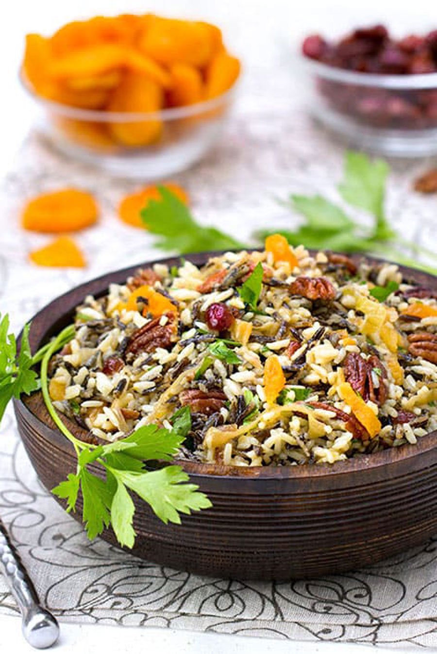 wild rice salad in a brown wooden bowl with a small bowl of dried apricots and a small bowl of dried cranberries in the background