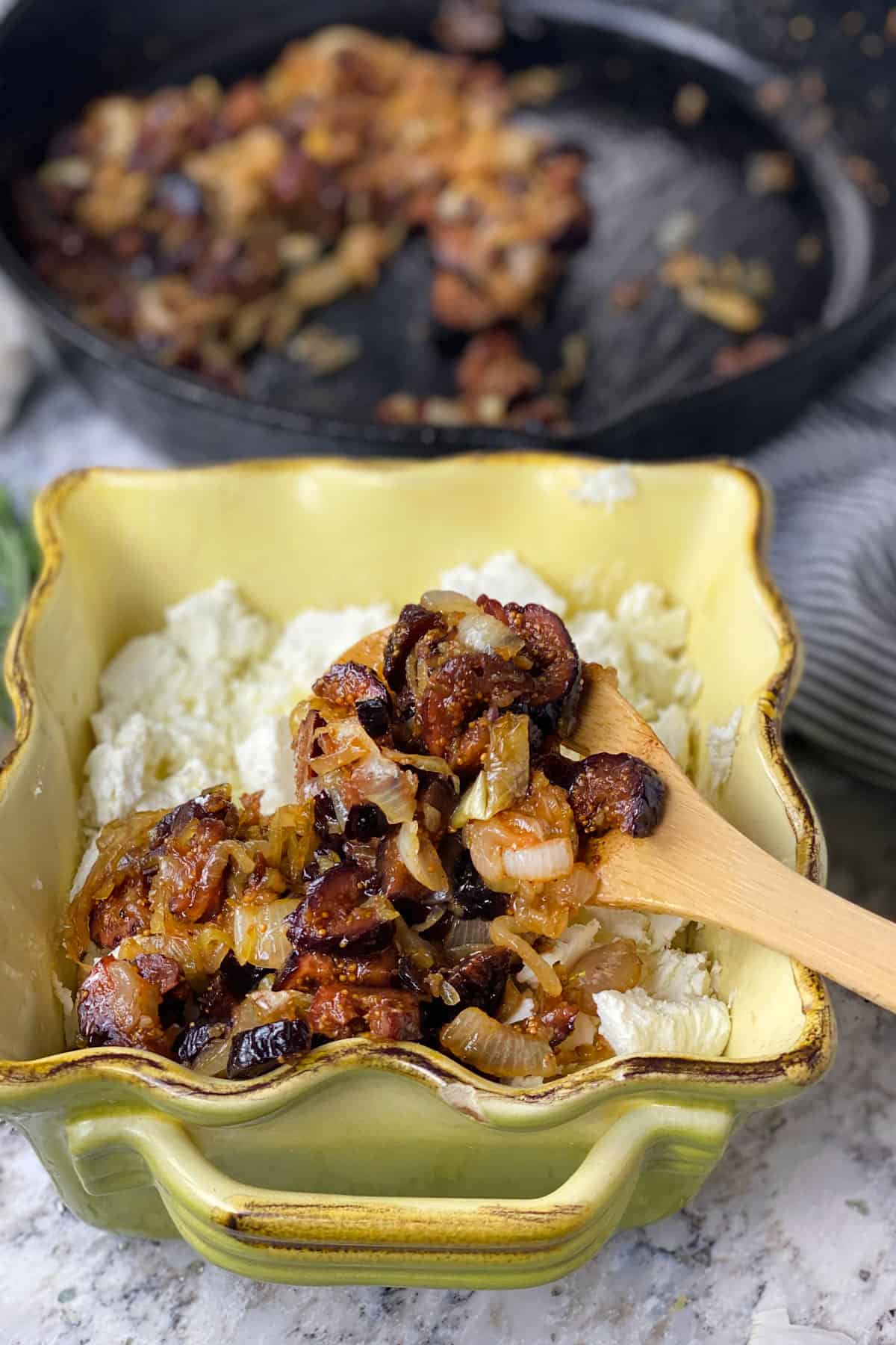 Fig and onions mixture being spooned on top of goat cheese, in a small square casserole.