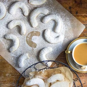 A dozen or so almond crescent cookies on a piece of white parchment plus a basket of crescent cookies and a cup of coffee to the side.