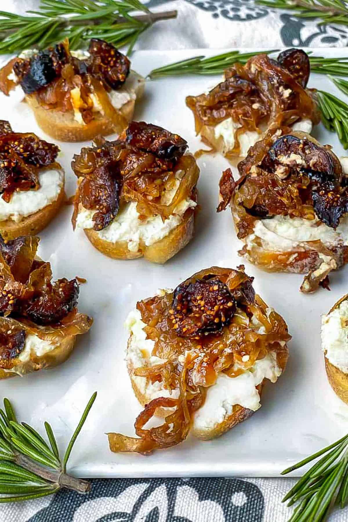 7 fig and goat cheese topped crostini on a square serving platter with sprigs of rosemary to garnish