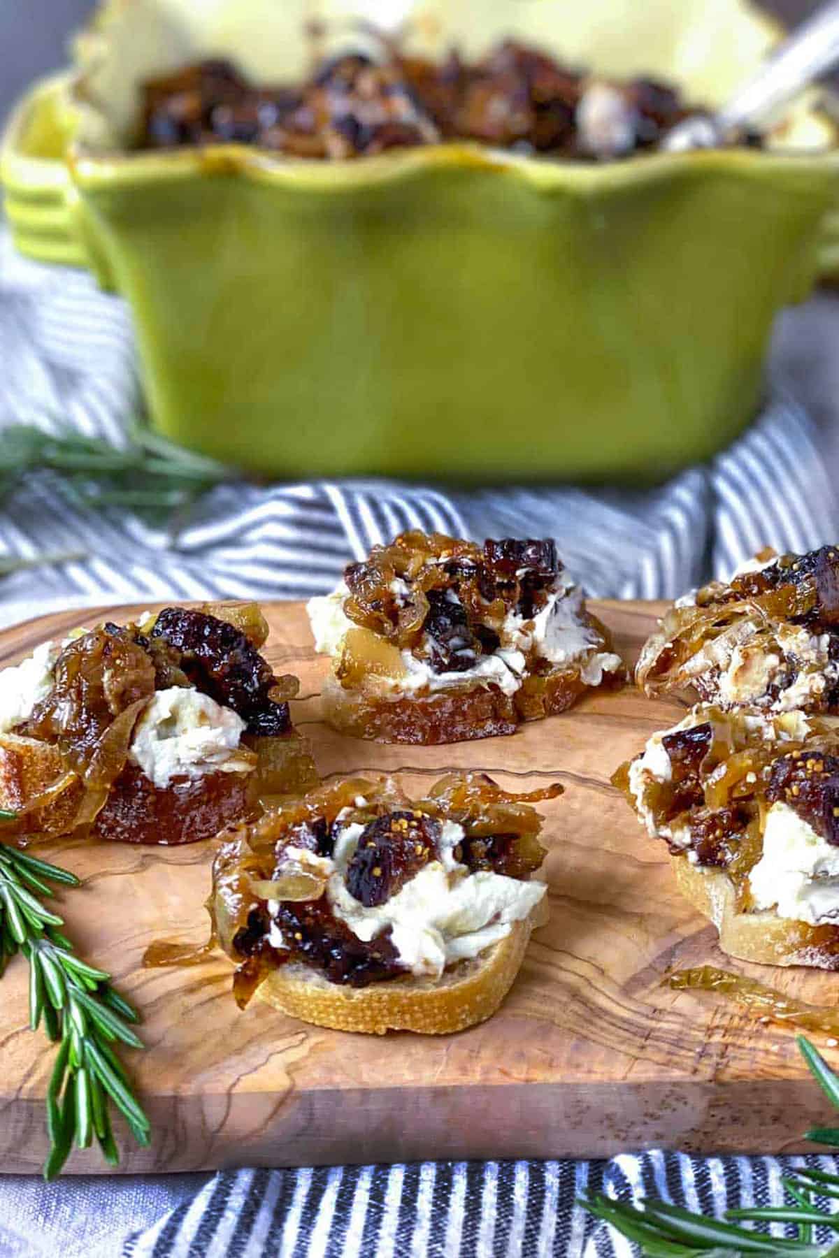 4 pieces of fig and goat cheese bruschetta on a wooden serving tray with a sprig of rosemary