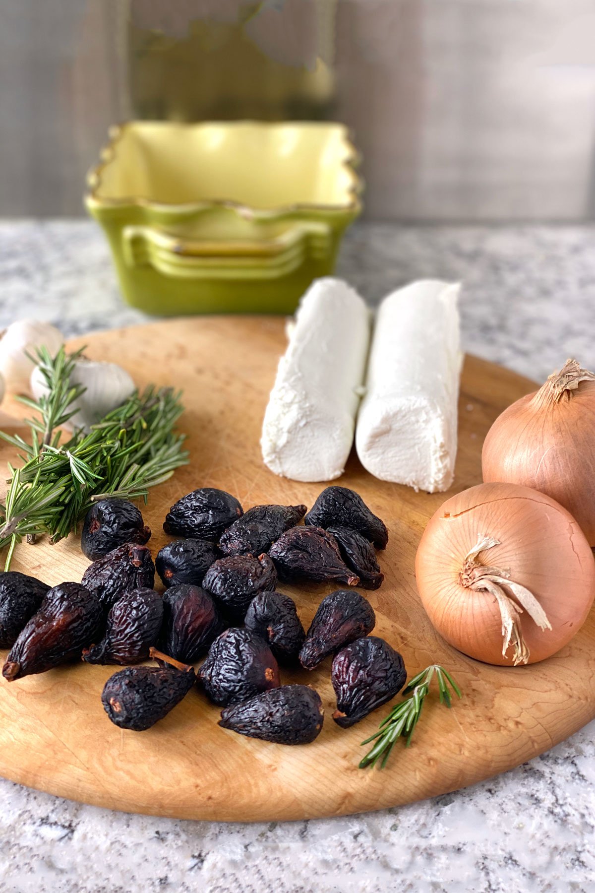 a dozen dried mission figs on a wooden cutting board along with 2 onions, 2 logs of goat cheese, a few rosemary sprigs and two bulbs of garlic