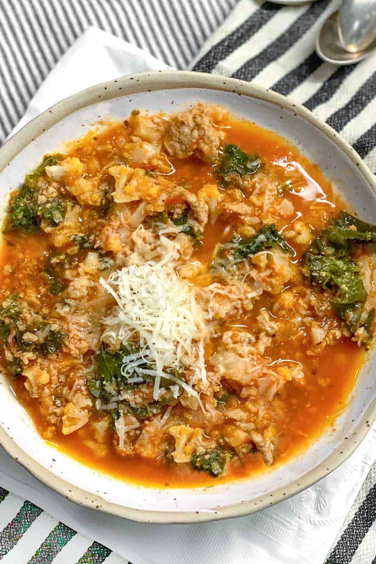Ceramic bowl filled with tomato based soup that has lots of ground turkey, chopped cauliflower and kale. Topped with grated parmesan.