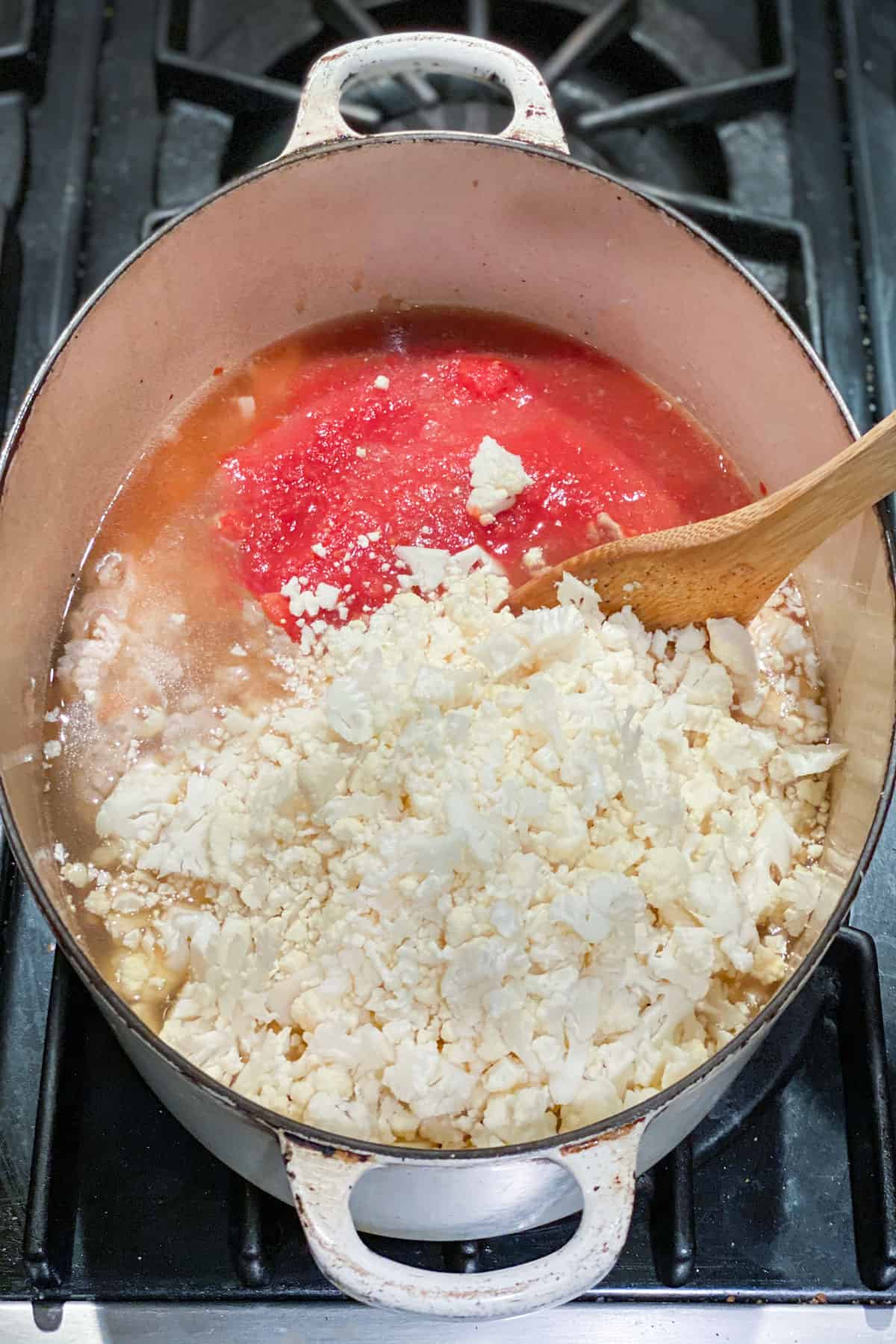 Raw chopped cauliflower and tomato puree being stirred with a wooden spoon, in a dutch oven.