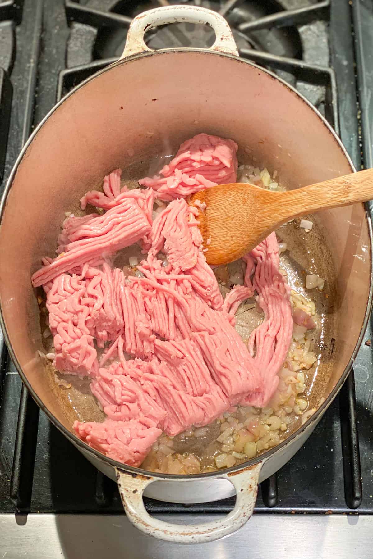 Raw ground turkey in an oval Dutch oven being stirred with a wooden spoon.