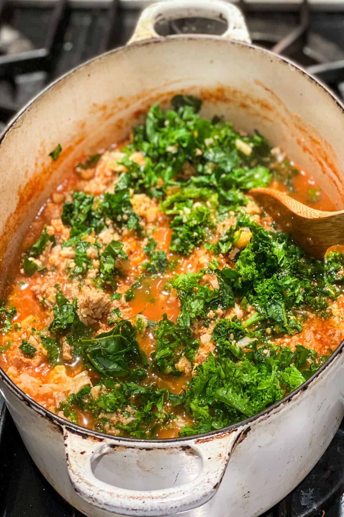Raw chopped curly kale being stirred into a pot of ground turkey soup, using a wooden spoon.
