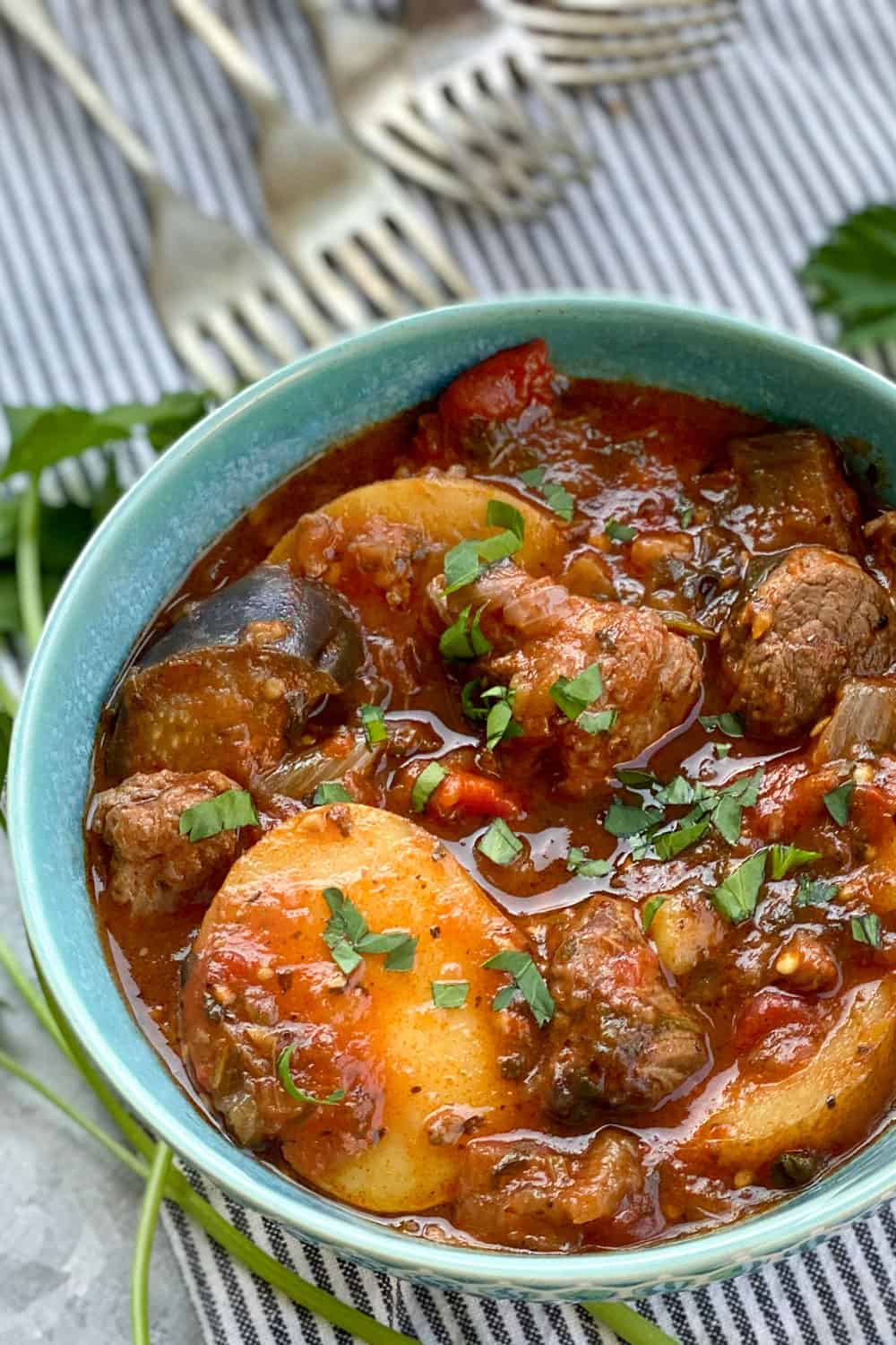 Georgian lamb stew called chanakhi, with potatoes in an aqua bowl on a striped dish cloth with 5 forks in the background