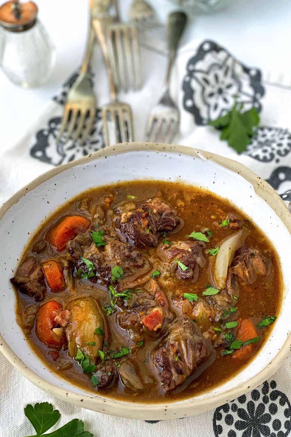 while bowl filled with beef bourguignon, 3 forks and a sprig of parsley in the background