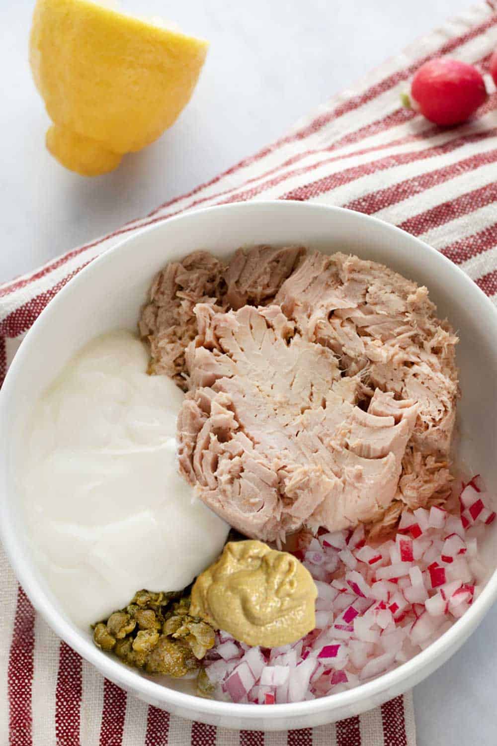 A large white bowl filled with ingredients for making healthy tuna salad: tuna, yogurt, mustard, chopped radishes and chopped red onions