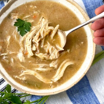 Creamy chicken soup with lots of shredded chicken in a white bowl with a leaf of parsley and a spoon